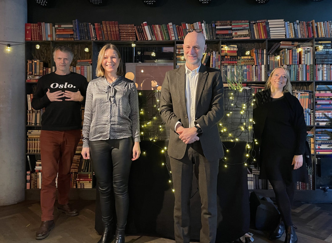 Erling Fossen, Matilda Vinje, Erling Dokk Holm og Maren Bjerkeng står på en scene og smiler mot kameraet. I bakgrunnen er en bokhylle og fin julebelysning.